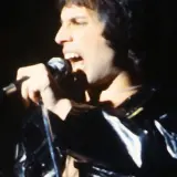 Freddie Mercury śpiewa do mikrofonu, ubrany w błyszczącą, czarną kurtkę. Jego usta są szeroko otwarte, a oczy zamknięte.