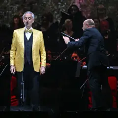 Andrea Bocelli w złotej marynarce śpiewa na scenie, dyrygent prowadzi orkiestrę. Koncert zapowiada się na niezapomniane przeżycie, jakiego można spodziewać się po Andrea Bocelli koncert 2026.