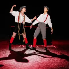 Dwie postacie w strojach z epoki, z szelkami i czapkami, na scenie. Jedna napina łydkę ferdydurke, druga ciągnie ją za szelki.