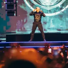 Pitbull Polska na scenie, w czarnej kurtce i spodniach, śpiewa do mikrofonu. Tłum w dole nagrywa koncert.