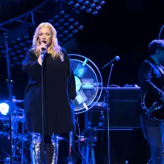 Wokalistka w czarnej sukience i błyszczących kozakach śpiewa do mikrofonu. Na scenie gra zespół, w tym gitarzysta i perkusista.