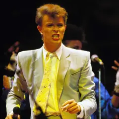 David Bowie na scenie podczas Live Aid, w jasnej marynarce i żółtym krawacie, śpiewa z pasją.