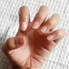Dłoń z naturalnymi paznokciami, lekko zgięta, jakby chciała powiedzieć "hold my hand". Tło białe, fakturowane.