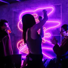 Trzy osoby bawią się przy energicznych piosenkach do tańca w klubie z neonowym oświetleniem.