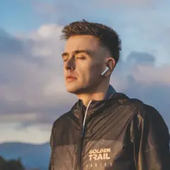 Mężczyzna w słuchawkach ANC, z zamkniętymi oczami, w kurtce z napisem "Golden Trail Series", na tle nieba o zachodzie słońca.