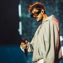 Taco Hemingway w okularach i szarej bluzie z kapturem, trzymający mikrofon. Fani czekają na koncert Taco Hemingway 2026.