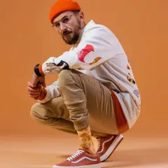 DJ BRK robię co mogę, w pomarańczowej czapce i okularach, kuca na pomarańczowym tle.
