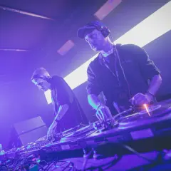 Dwóch DJ-ów w słuchawkach, tworzących muzykę techno. Jeden z nich ma na szyi identyfikator "ARTIST".