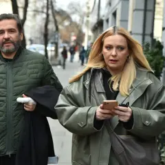 Paweł Krawczyk i kobieta idą ulicą. On ma zieloną, pikowaną kurtkę i czarny płaszcz w ręku. Ona patrzy w telefon.