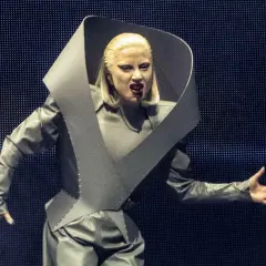 Lady Gaga w szarym, futurystycznym stroju na koncercie w Polsce, z charakterystycznym kołnierzem i makijażem.