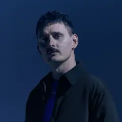 Teddy Swims z wąsami i w ciemnej kurtce na granatowym tle.