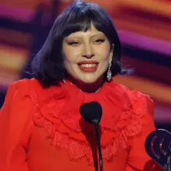 Lady Gaga w czerwonej sukni z falbankami, uśmiechnięta na scenie podczas koncertu. W ręku trzyma nagrodę.