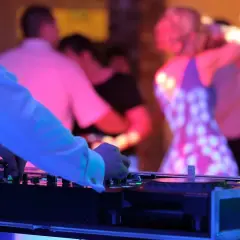 DJ przy konsoli, w tle tańczący ludzie na weselu w kolorowych światłach.