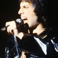 Freddie Mercury śpiewa do mikrofonu, ubrany w błyszczącą, czarną kurtkę. Jego usta są szeroko otwarte, a oczy zamknięte.