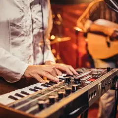 Muzyk gra na keyboardzie, tworząc tło dla piosenek do śpiewania. W tle widać gitarzystę.