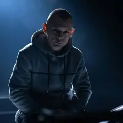 Młody raper w szarej bluzie z kapturem, oświetlony od tyłu, wpatruje się w obiektyw.