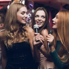 Trzy kobiety śpiewają do mikrofonów, bawiąc się przy najlepszych karaoke. W tle widać innych ludzi na imprezie.
