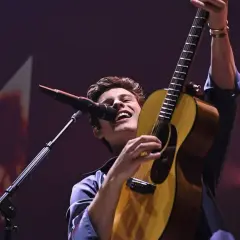 Shawn Mendes na koncercie, śpiewa i gra na gitarze, wznosząc ją w górę.