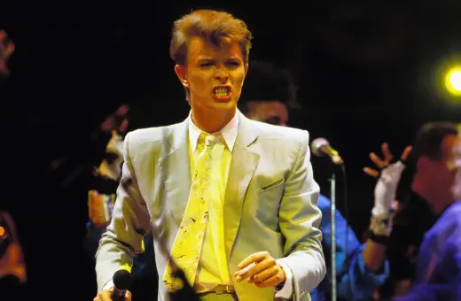 David Bowie na scenie podczas Live Aid, w jasnej marynarce i żółtym krawacie, śpiewa z pasją.