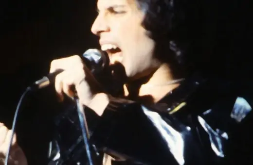 Freddie Mercury śpiewa do mikrofonu, ubrany w błyszczącą, czarną kurtkę. Jego usta są szeroko otwarte, a oczy zamknięte.