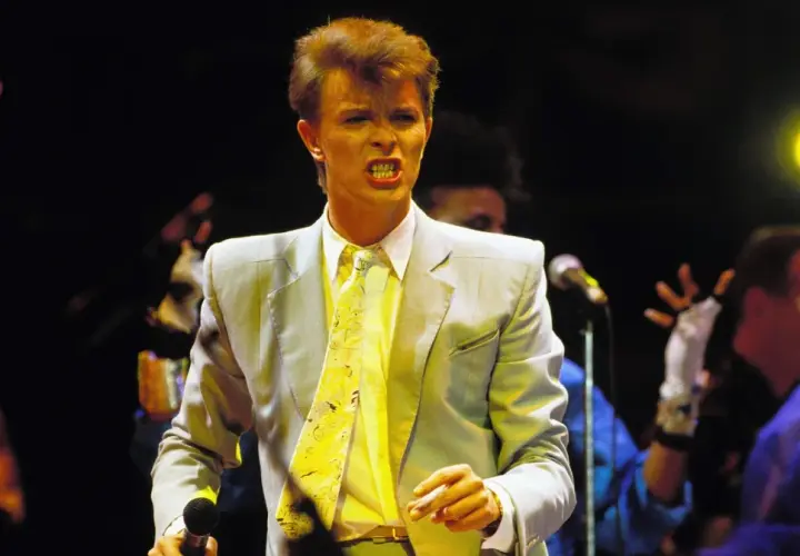 David Bowie na scenie podczas Live Aid, w jasnej marynarce i żółtym krawacie, śpiewa z pasją.