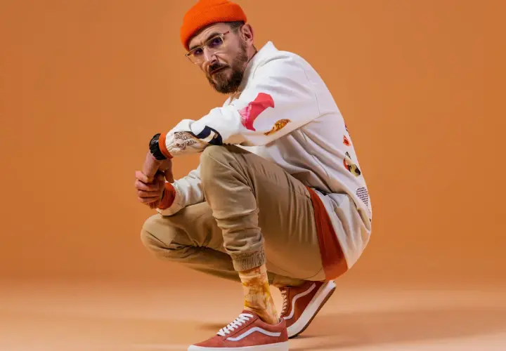 DJ BRK robię co mogę, w pomarańczowej czapce i okularach, kuca na pomarańczowym tle.