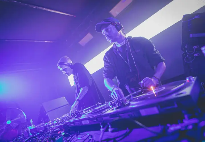 Dwóch DJ-ów w słuchawkach, tworzących muzykę techno. Jeden z nich ma na szyi identyfikator "ARTIST".