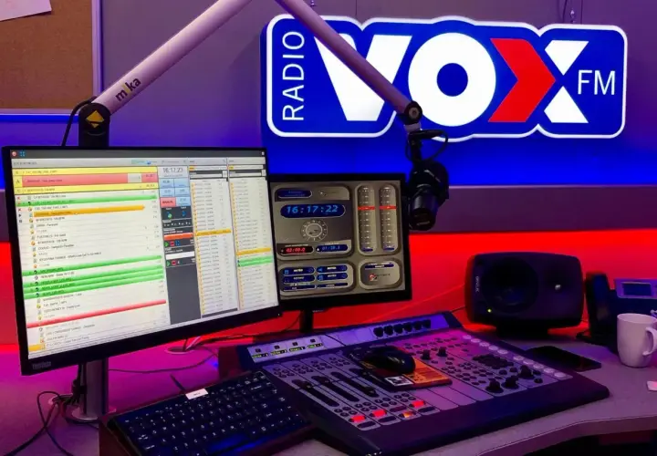 Studio radiowe VOX FM Warszawa. Komputery z listą utworów, mikser, mikrofon i logo stacji.