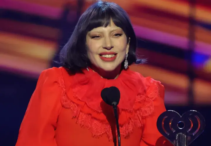 Lady Gaga w czerwonej sukni z falbankami, uśmiechnięta na scenie podczas koncertu. W ręku trzyma nagrodę.