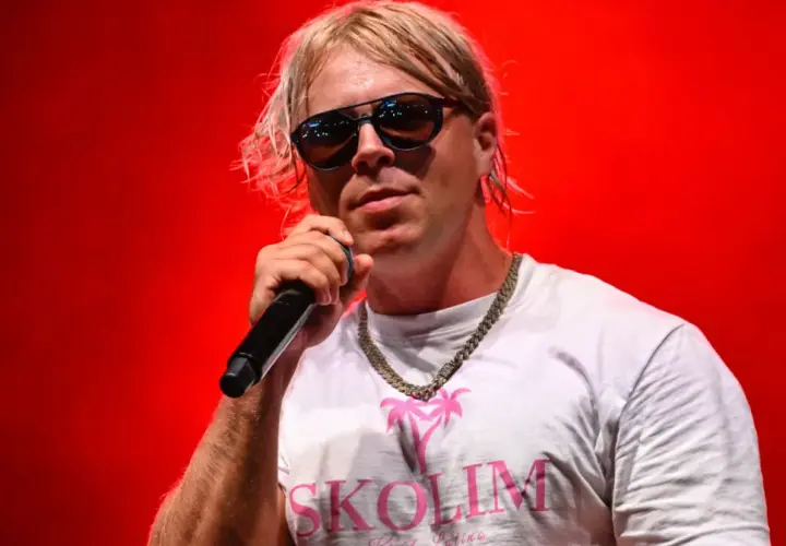 Skolim w okularach i białej koszulce z napisem "SKOLIM" śpiewa na czerwonym tle. Zastanawiasz się, ile Skolim bierze za koncert?