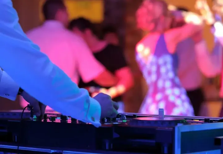 DJ przy konsoli, w tle tańczący ludzie na weselu w kolorowych światłach.