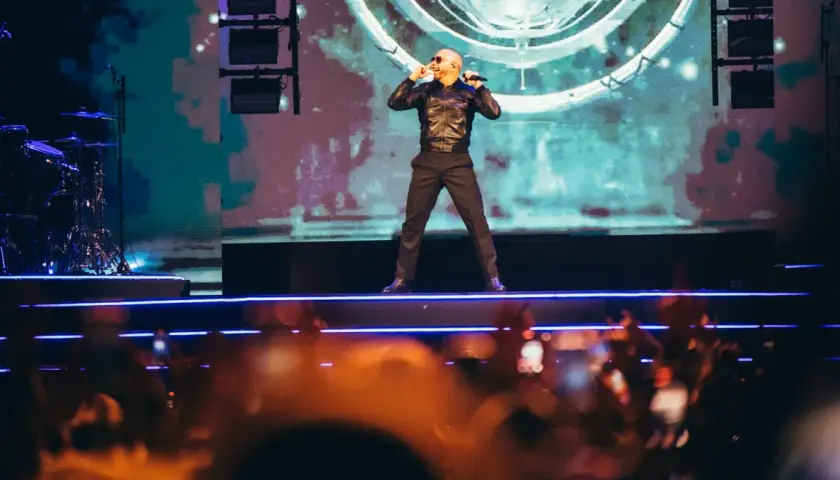 Pitbull Polska na scenie, w czarnej kurtce i spodniach, śpiewa do mikrofonu. Tłum w dole nagrywa koncert.