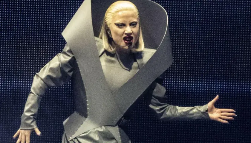 Lady Gaga w szarym, futurystycznym stroju na koncercie w Polsce, z charakterystycznym kołnierzem i makijażem.