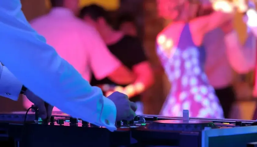 DJ przy konsoli, w tle tańczący ludzie na weselu w kolorowych światłach.