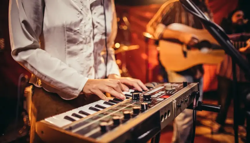 Muzyk gra na keyboardzie, tworząc tło dla piosenek do śpiewania. W tle widać gitarzystę.