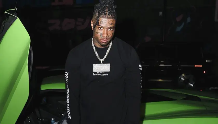 Raper z tatuażami na twarzy, znany jako blueface, pozuje obok jaskrawozielonego samochodu sportowego.