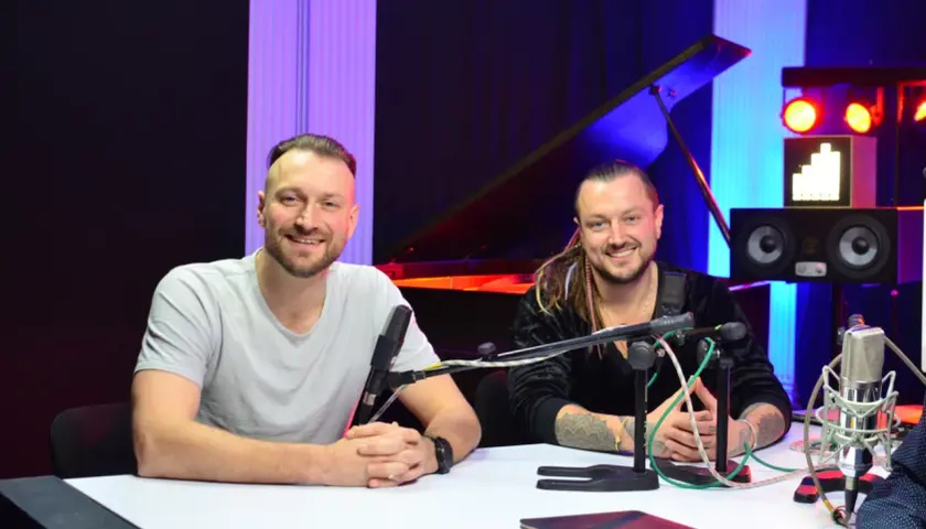 Alek Baron i jego kolega siedzą przy stole w studiu, gotowi do nagrania.