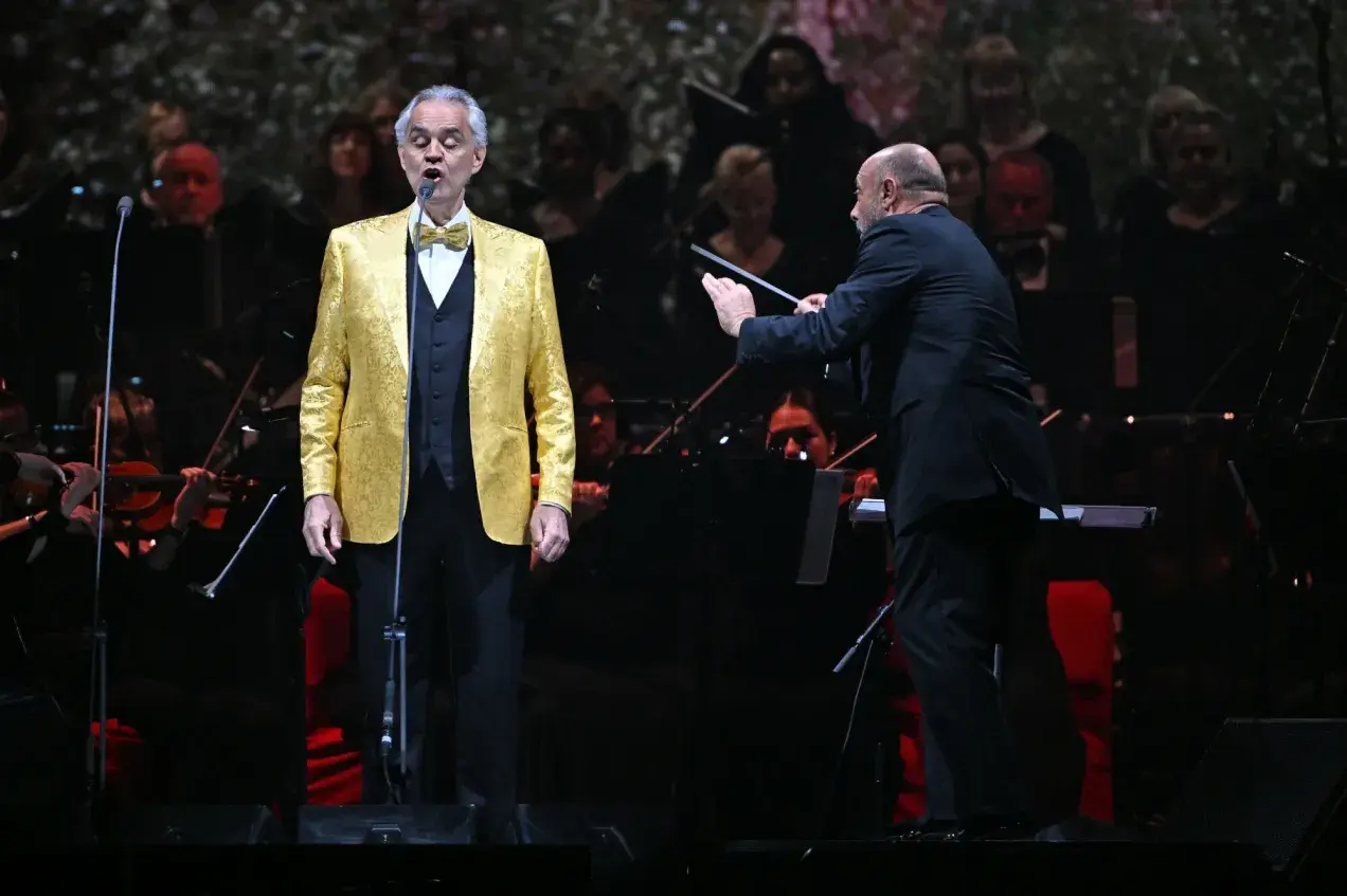 Andrea Bocelli w złotej marynarce śpiewa na scenie, dyrygent prowadzi orkiestrę. Koncert zapowiada się na niezapomniane przeżycie, jakiego można spodziewać się po Andrea Bocelli koncert 2026.