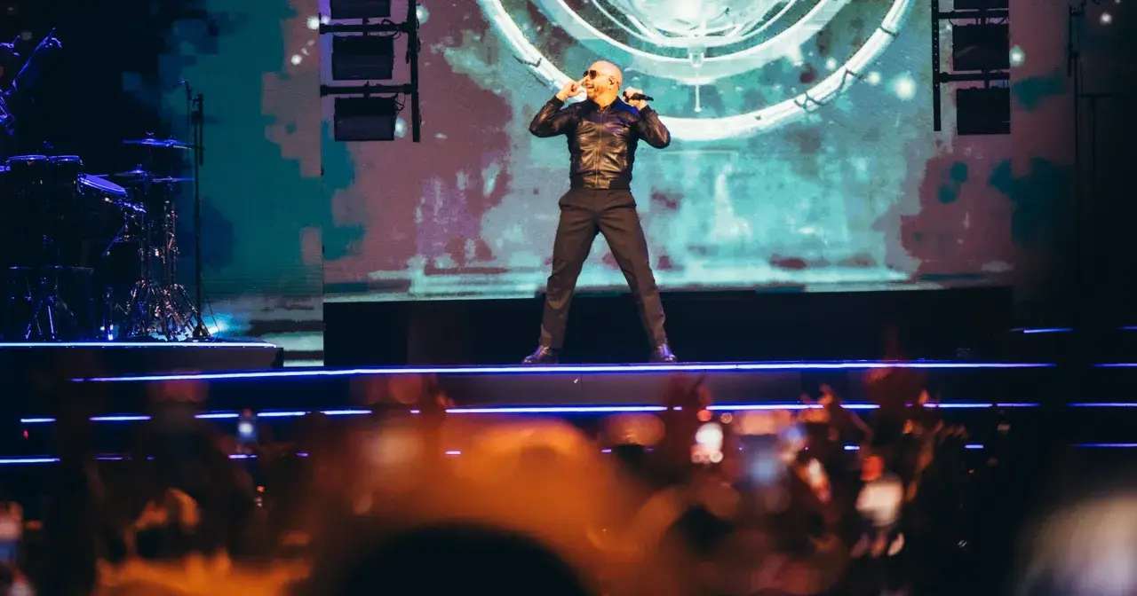 Pitbull Polska na scenie, w czarnej kurtce i spodniach, śpiewa do mikrofonu. Tłum w dole nagrywa koncert.