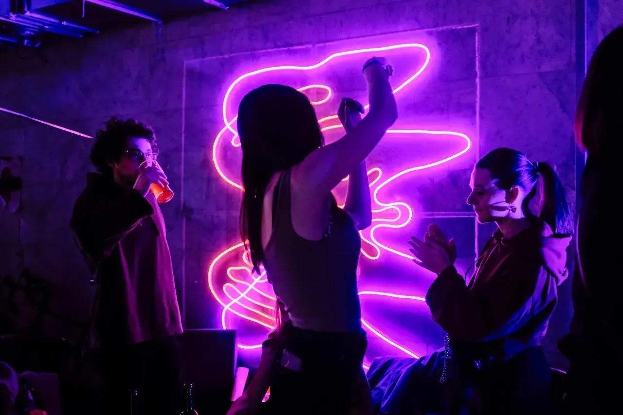 Trzy osoby bawią się przy energicznych piosenkach do tańca w klubie z neonowym oświetleniem.