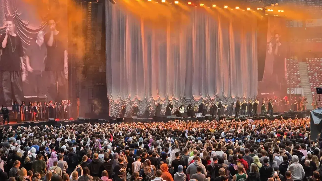 Ogromny tłum na quebo koncercie, fani nagrywają występ na swoich telefonach. Na scenie artyści i muzycy.