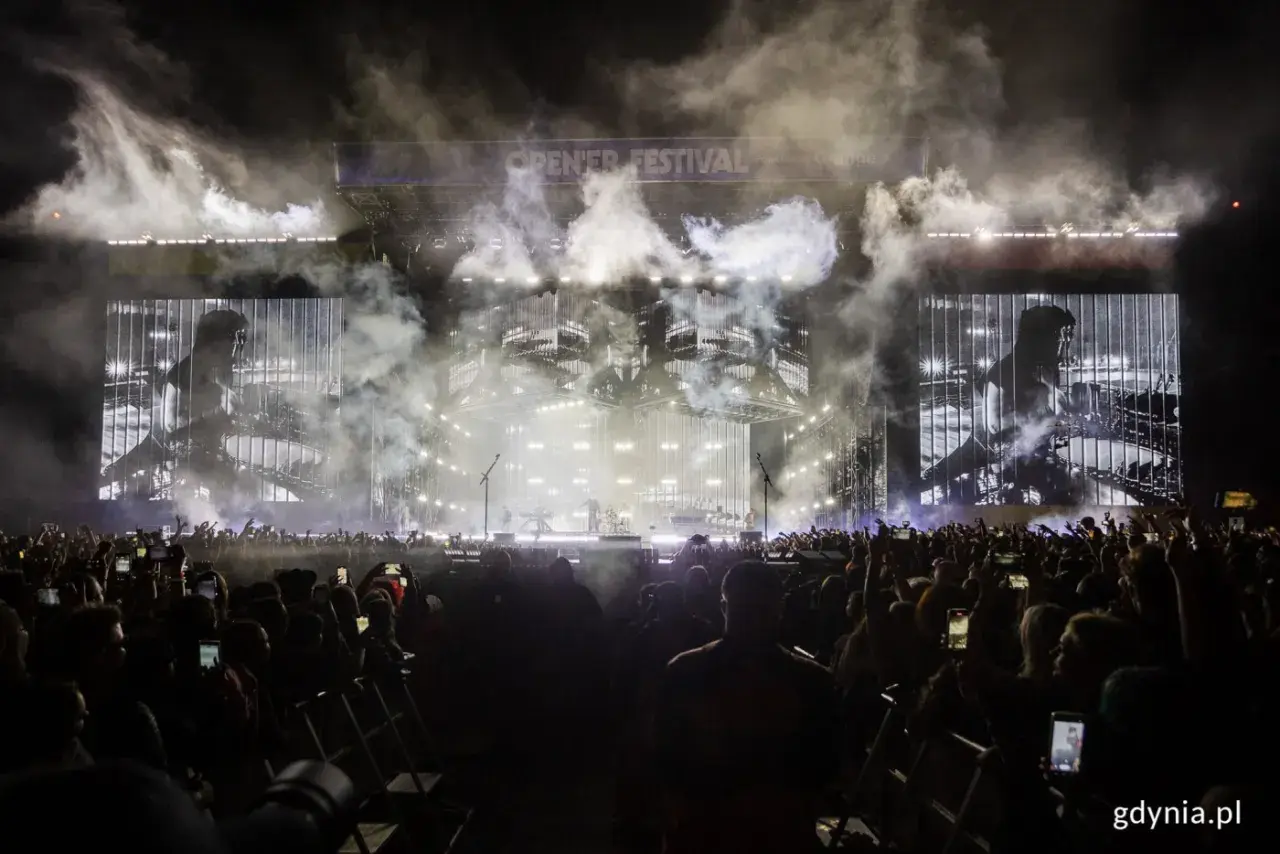 Koncert Linkin Park Polska na Open'er Festival. Tłum ludzi z telefonami, dym i światła na scenie.
