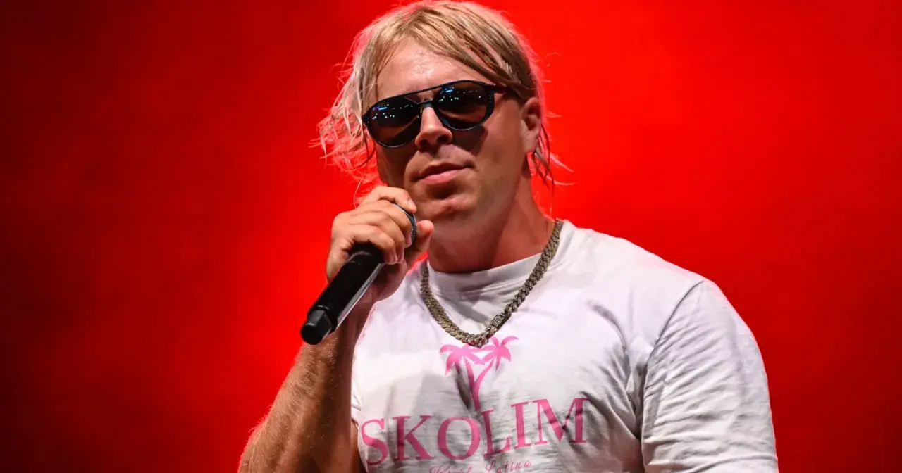 Skolim w okularach i białej koszulce z napisem "SKOLIM" śpiewa na czerwonym tle. Zastanawiasz się, ile Skolim bierze za koncert?
