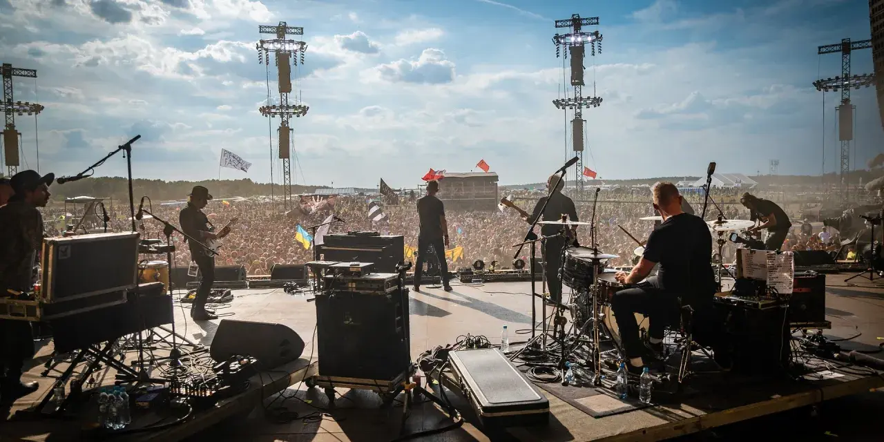 Zespół gra na scenie festiwalowej, tłum szaleje. W tle widać flagi, w tym z napisem "Sztywny Pal Azji utwory".
