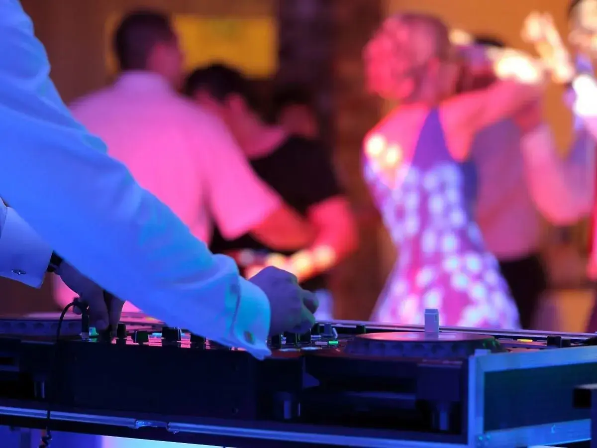 DJ przy konsoli, w tle tańczący ludzie na weselu w kolorowych światłach.