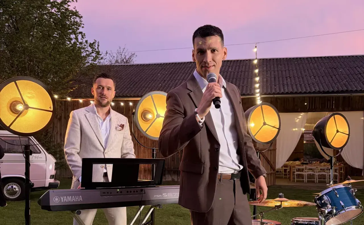 Zespół Defis gotowy do występu na świeżym powietrzu. Wokalista z mikrofonem, klawiszowiec przy keyboardzie, a w tle światła i budynek.