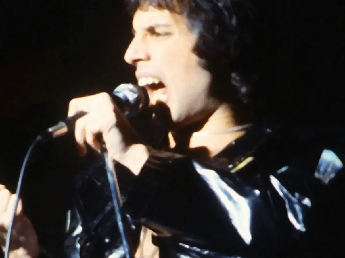 Freddie Mercury śpiewa do mikrofonu, ubrany w błyszczącą, czarną kurtkę. Jego usta są szeroko otwarte, a oczy zamknięte.