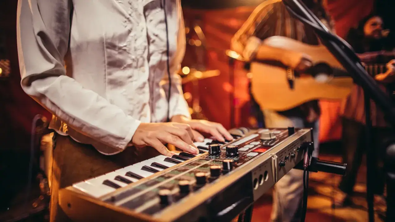 Muzyk gra na keyboardzie, tworząc tło dla piosenek do śpiewania. W tle widać gitarzystę.