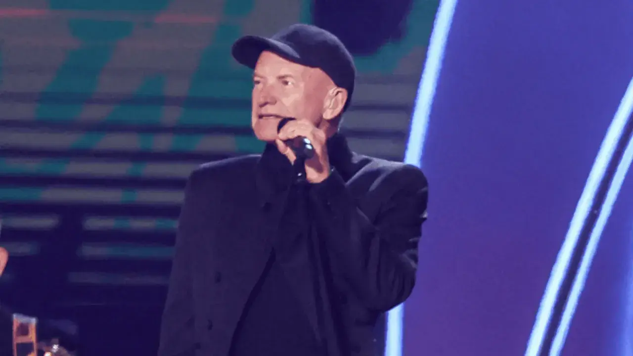 Sting w czapce i czarnym stroju śpiewa do mikrofonu. Ciekawe, ile Sting bierze za koncert, gdy występuje na tak dużej scenie.