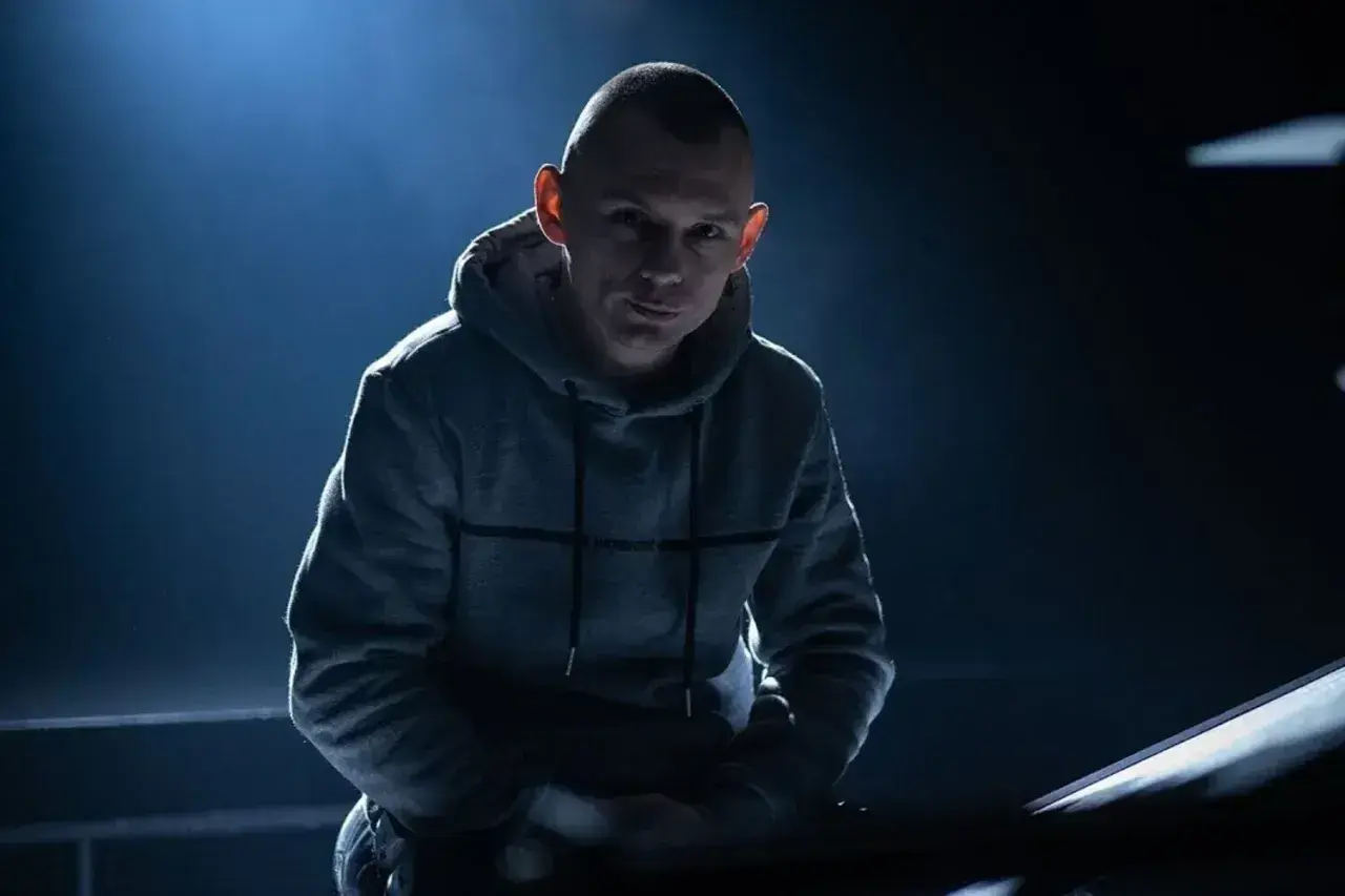 Młody raper w szarej bluzie z kapturem, oświetlony od tyłu, wpatruje się w obiektyw.